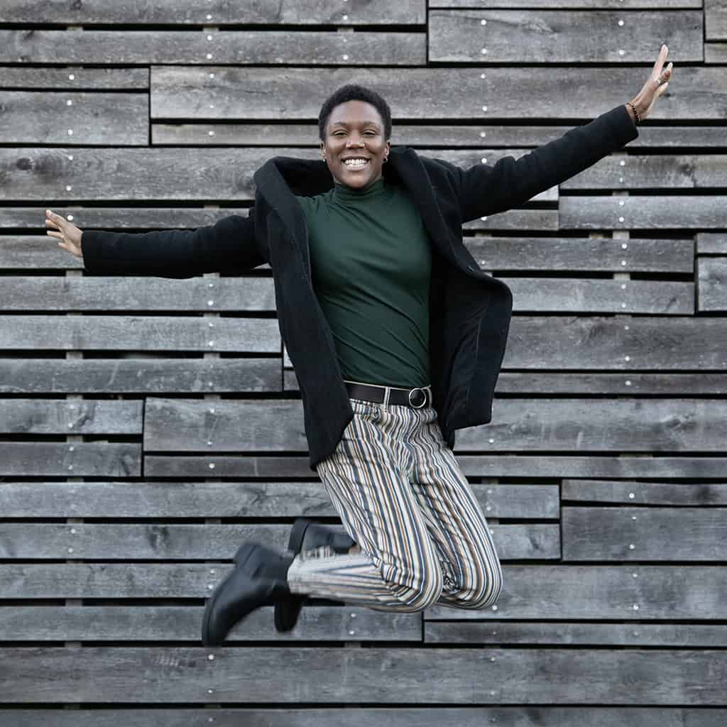 Ambre de vie sautant de manière énergique et avec le sourire devant un mur en bois. Elle porte un long manteau noir, un col roulé vert et un pantalon rayé